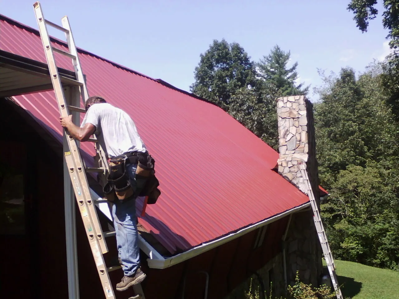 Roofer on a ladder beside a red metal roof and stone chimney — Soffit & Fascia Repair by Roofing Brothers in Franklin Lakes